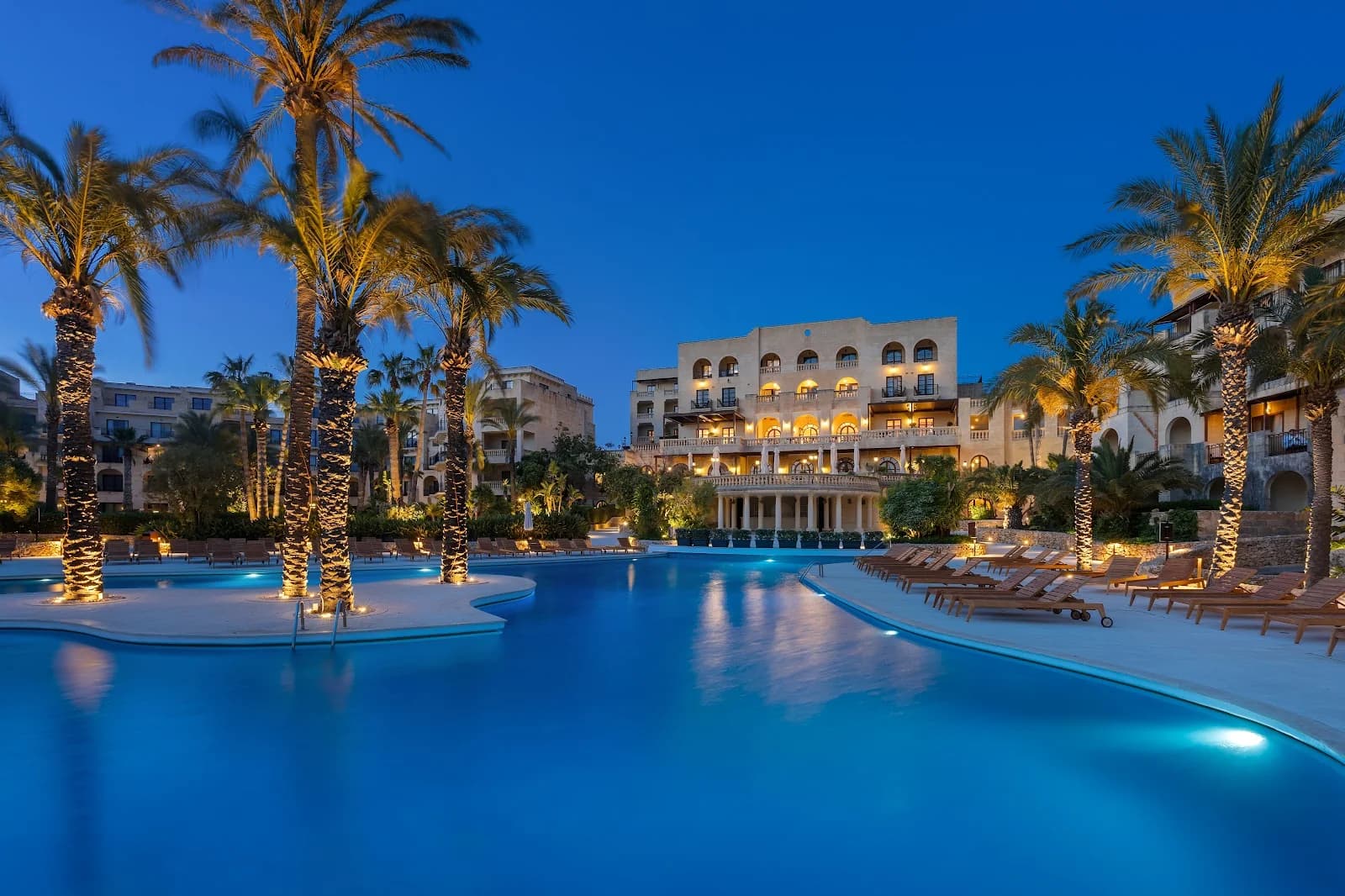 Paved entrance path to a luxury spa in Gozo, Malta, lined with tall palm trees, vibrant pink bougainvillea, and citrus.